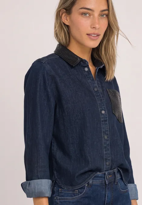 Jonge vrouw in een donkerblauwe denimblouse, mouwen omgeslagen, neutrale achtergrond.