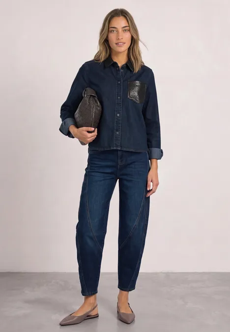Vrouw in donker denim overhemd en -broek, met een bruine clutch, platte schoenen, tegen een neutrale achtergrond.