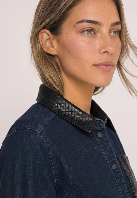Een vrouw met lichtbruin haar draagt spijkerjack met zwarte leren kraag en kijkt zijdelings voor neutrale achtergrond.