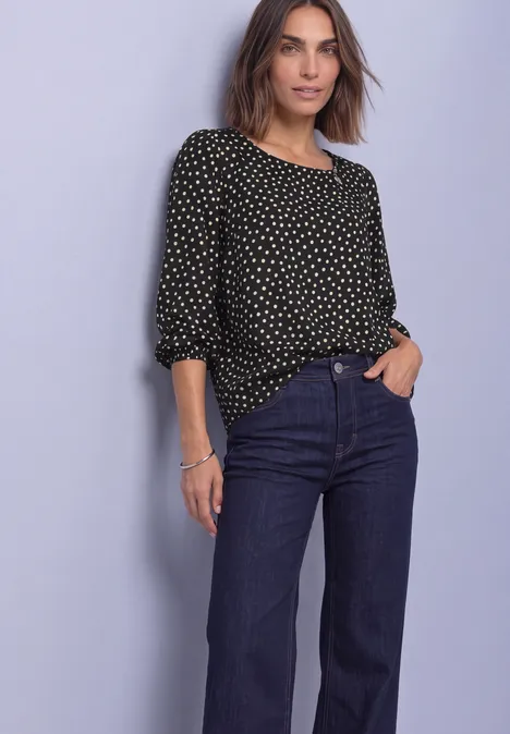 Frau in schwarzer Bluse mit weißen Punkten und weiten, dunkelblauen Jeans vor hellem Hintergrund.