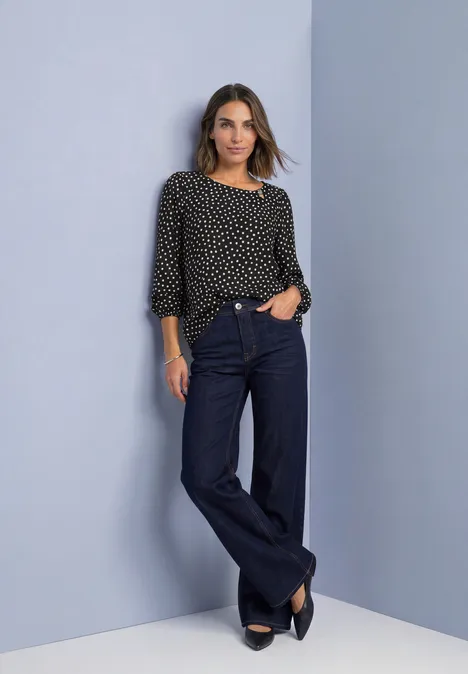 Frau in schwarzer Polka-Dot-Bluse und weiten Jeans, posiert lässig vor neutralem Hintergrund.
