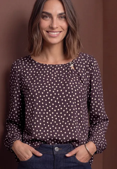 Chemisier à col rond et détail à boucles berry plum Lächelnde Frau trägt braune Bluse mit beigen Punkten und posiert vor schlichtem braunem Hintergrund.