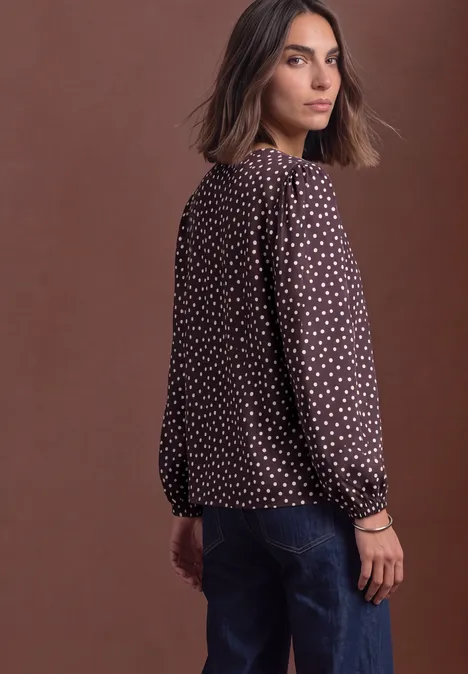 Chemisier à col rond et détail à boucles berry plum Eine Frau in brauner, weißgepunkteter Bluse und Jeans vor braunem Hintergrund, sie schaut über die Schulter.