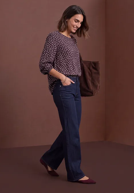 Chemisier à col rond et détail à boucles berry plum Frau in weiten Jeans und gepunkteter Bluse geht lächelnd vor braunem Hintergrund.