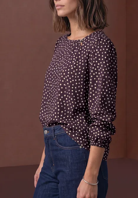 Chemisier à col rond et détail à boucles berry plum Eine Frau trägt eine dunkel gepunktete Bluse, blaue Jeans und ein Armband vor braunem Hintergrund.