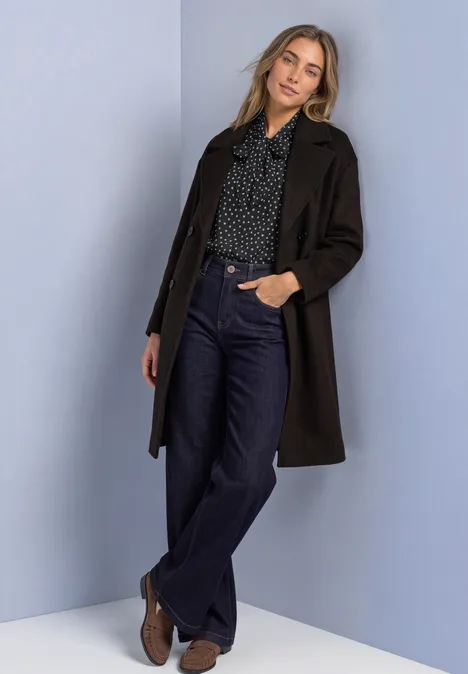 Frau in langem dunkelbraunen Mantel, gepunkteter Bluse, weiten Jeans und Stiefeletten vor schlichtem Hintergrund.
