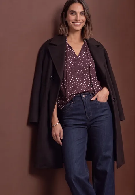 Haut-blouse en chiffon avec nœud décoratif berry plum Femme souriante en manteau sombre, chemisier violet à pois et jean bleu, posant devant un fond marron.