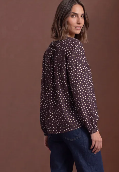 Eine Frau in seitlicher Pose trägt eine braune, gepunktete Bluse und Jeans vor braunem Hintergrund.