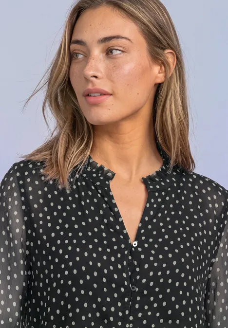Junge Frau in schwarzer, gepunkteter Chiffonbluse mit Rüschenkragen und transparenten Ärmeln.