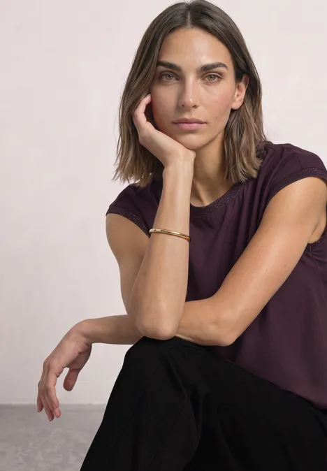 Blouse courtes manches à col rond et bandes pailletés berry plum Une femme aux cheveux bruns porte un t-shirt violet, appuie sa tête sur sa main et regarde l'objectif.