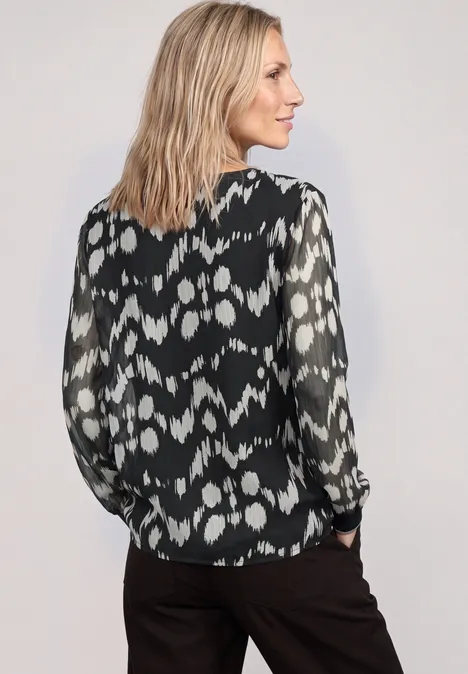 Frau in schwarzer Bluse mit weißem Muster, schaut seitlich. Schulterlange blonde Haare