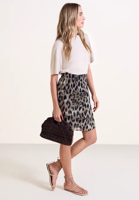 Frau im weißen Top mit Leopardenmuster-Rock, trägt gesträppte Sandalen und hält eine geflochtene Clutch.