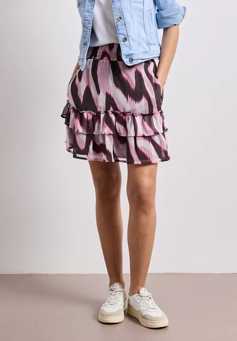 Une femme porte une jupe courte à volants rose et noire à motif, associée à une veste en jean et des baskets blanches.