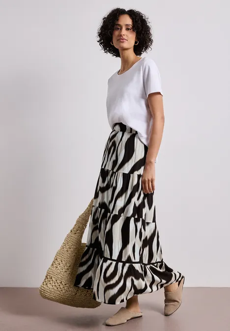 Frau in weißem T-Shirt, langer Zebra-Muster-Rock, Strohtasche und beigen Mules vor neutralem Hintergrund.