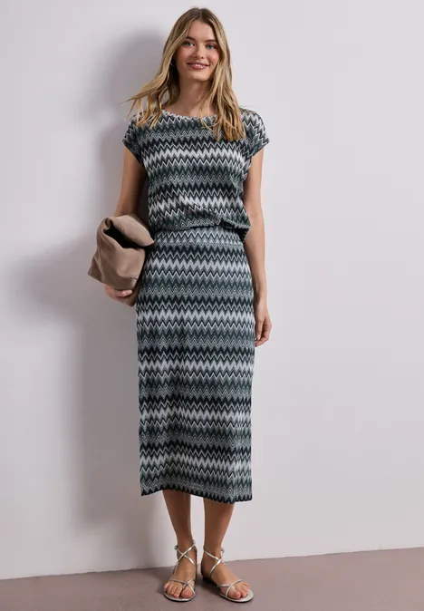Vrouw in een midi-jurk met korte mouwen en zigzagprint, platte sandalen en clutch in neutrale kleur.