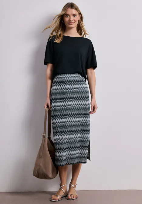 Een vrouw draagt een zwart T-shirt, een midi-rok met patroon, sandalen en houdt een grote tas.