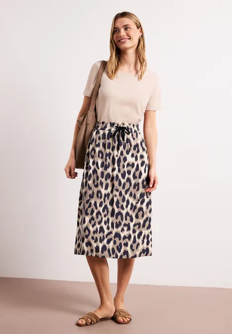 Vrouw in beige top, midi rok met luipaardprint, sandaaltjes en schoudertas, tegen witte achtergrond.