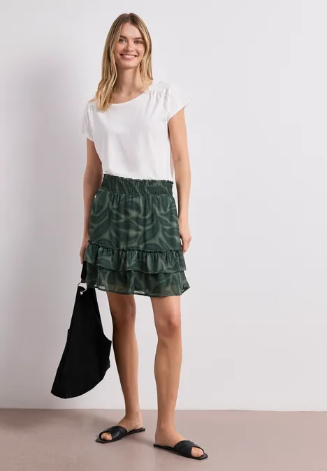 Frau in weißem T-Shirt, grün gemustertem Stufenrock, schwarzen Slides und schwarzer Tasche vor weißem Hintergrund.