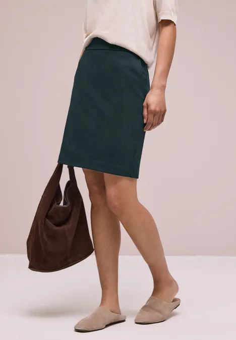 Vrouw in witte blouse en groene rok met bruine tas en beige instappers voor een neutrale achtergrond.