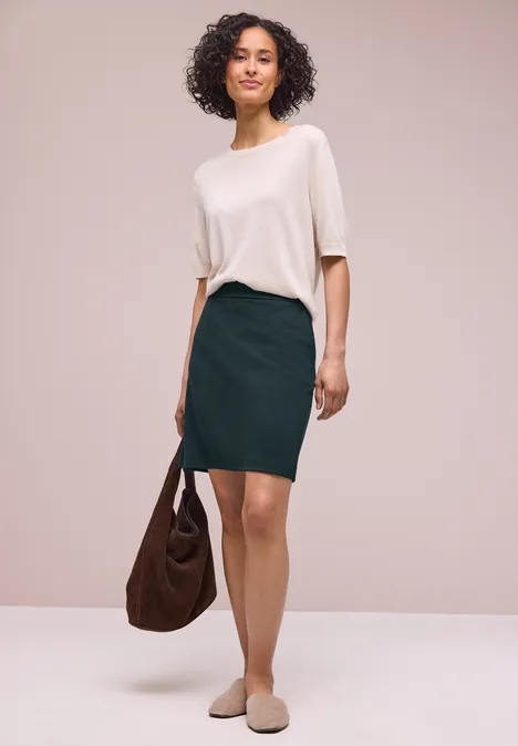 Vrouw met krullend haar draagt witte top, groene kokerrok, beige muiltjes en houdt een bruine tas vast.