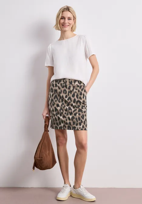 Frau trägt weißen T-Shirt, Leopardenmuster-Rock, weiße Sneaker und hält eine braune Tasche.