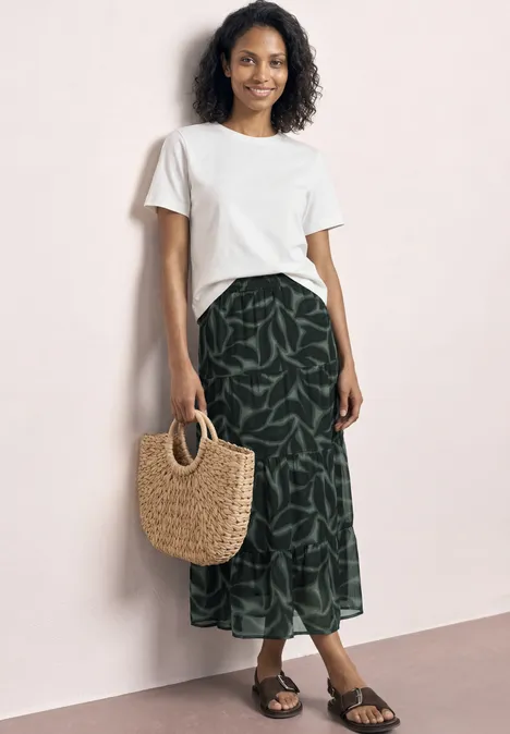 Vrouw voor roze muur met wit shirt, groene maxirok met bladprint en strooien tas.