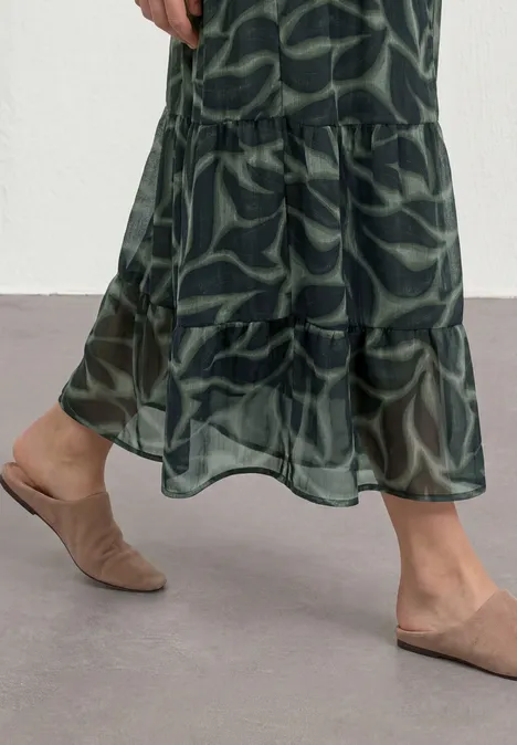 Frau trägt grünen Maxirock mit Blattmuster und transparenter Stufe, dazu beige Mules auf grauem Boden.
