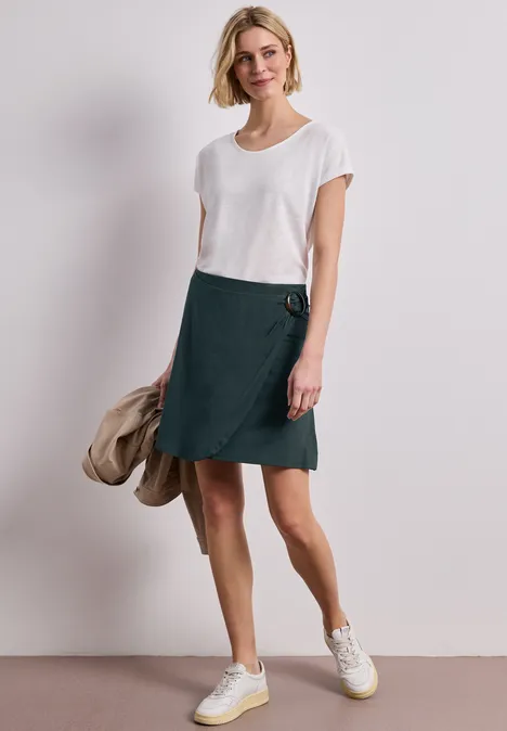 Frau trägt weißes T-Shirt mit grünem Wickelrock und Sneakers, hält eine Jacke in der Hand.