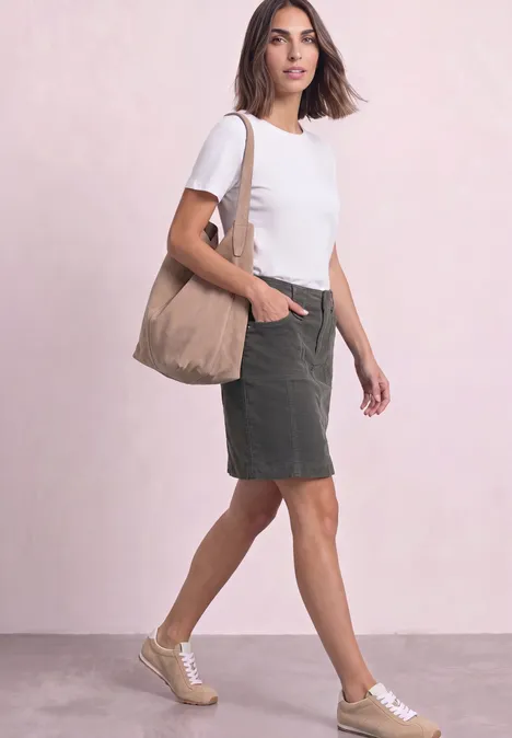 Vrouw in wit t-shirt, groene rok en sneakers, met een beige tas voor een roze achtergrond.
