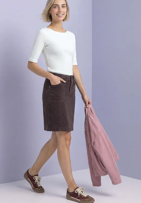 Vrouw in wit 3/4-shirt en bruine rok, draagt sneakers, houdt een roze jasje vast, tegen een lichtpaarse achtergrond.