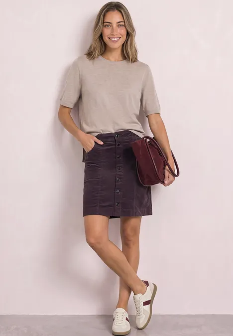 Jonge vrouw in beige top, paarse rok met knopen, witte sneakers en een bordeaux schoudertas, glimlachend.