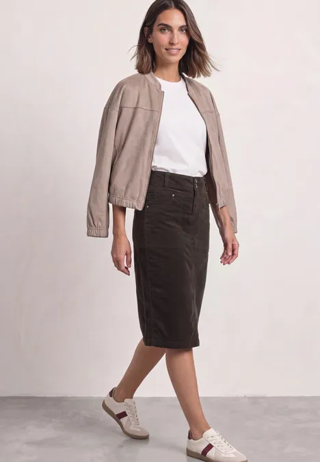 Une femme porte un t-shirt blanc, une veste en cuir beige, une jupe crayon noire et des baskets blanches.