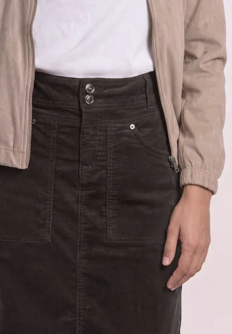 Une personne porte un t-shirt blanc, une veste beige et une jupe en velours côtelé noire avec poches.