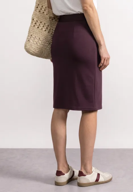 Rückansicht einer Person in einem violetten knielangen Rock, weißen Sneakers und Tragetasche.