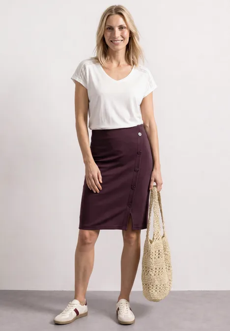 Eine Frau in weißem T-Shirt, bordeauxfarbener Knopfrock, weiße Sneaker und netzartige Tasche