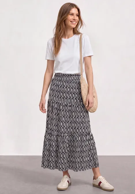 Maxi-Rock mit Volants und Print clay sand Frau trägt weißes T-Shirt, gemusterten Maxi-Rock, helle Schnürschuhe und geflochtene Schultertasche.