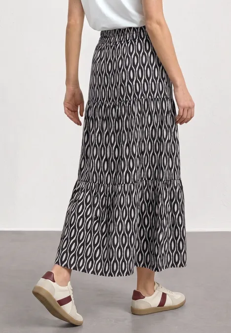 Maxi-Rock mit Volants und Print clay sand Frau im weißen T-Shirt trägt einen langen, schwarz-weißen Rock mit geometrischem Muster und weiße Sneaker.