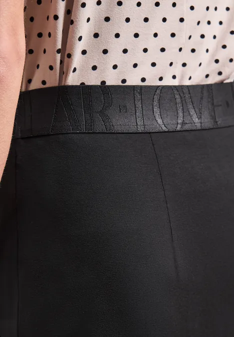 Midirok van tricot in effen kleur Black Vrouw draagt wit topje met zwarte stippen en een zwarte broek met geperst logo in de taille.