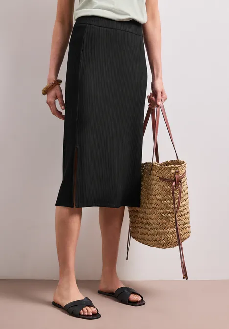 Geribde midirok met elastische tailleband Black Persoon draagt een zwarte midirok, zwarte sandalen en een gevlochten strozak.