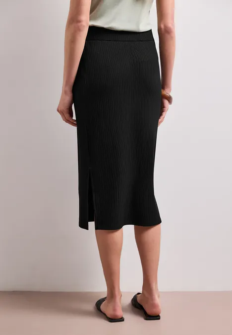 Geribde midirok met elastische tailleband Black Achteraanzicht van een zwarte midi-rok met zijsplit en zwarte slippers voor een neutrale achtergrond.