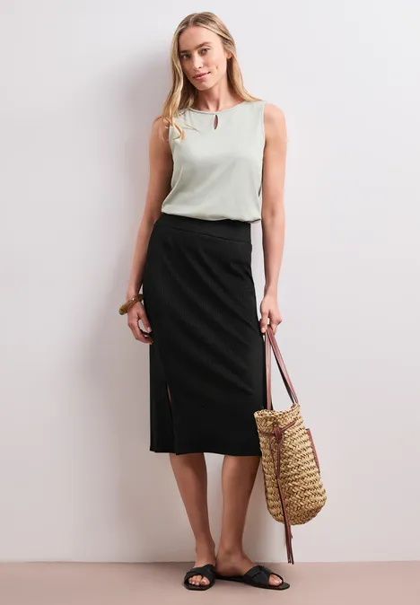 Geribde midirok met elastische tailleband Black Vrouw in wit mouwloos topje, zwarte midirok met split, zwarte sandalen en rieten tas.