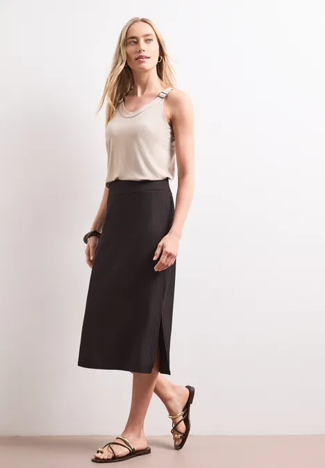 Geribde midirok met elastische tailleband espresso brown Vrouw met wit topje, zwarte midi-rok en sandalen voor een witte achtergrond.