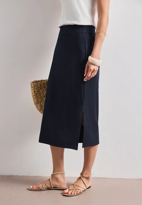 Frau in weißem Top und blauem Midi-Rock mit Schlitz, Strohtasche und flachen Sandalen.