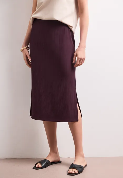 Geribde midirok met elastische tailleband rustic rouge Een aansluitende midi-rok in donkerpaars met zijsplitten, gecombineerd met een beige top en zwarte sandalen.