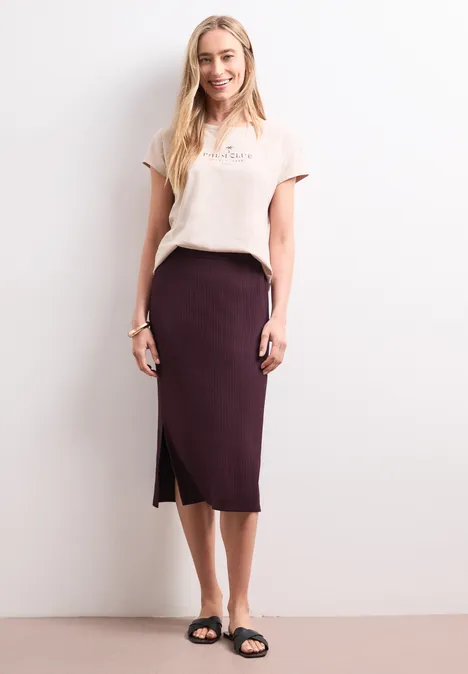 Geribde midirok met elastische tailleband rustic rouge Vrouw in beige t-shirt, donkerpaarse midirok met split en zwarte sandalen tegen witte achtergrond.