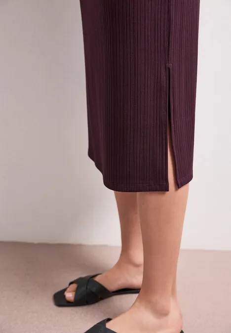 Geribde midirok met elastische tailleband rustic rouge Zwarte sandalen met geknoopte bandjes, bordeaux geribde midijurk met zijsplit op beige achtergrond.