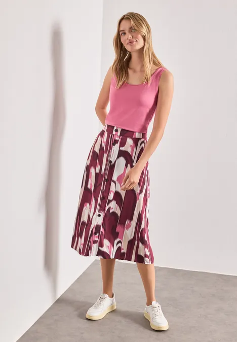 Eine Frau in rosa Top, gemustertem Midirock und weißen Sneakern steht lässig vor einer weißen Wand.