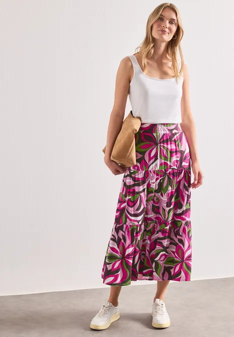 Maxi-Rock mit Volants und Print magenta dream Frau in weißem Top, knöchellangem Rock mit pink-grünem Blumenmuster, weißen Sneakern und beiger Tasche.