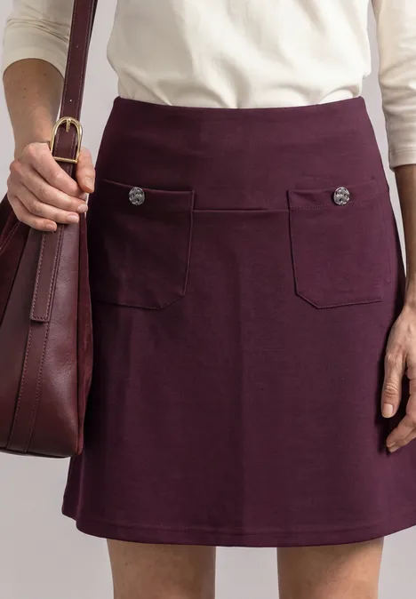 Dunkelvioletter Damenrock mit zwei aufgesetzten Taschen und Zierknöpfen, kombiniert mit cremefarbenem Oberteil.
