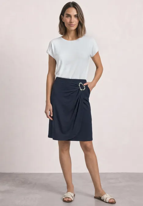 Vrouw in wit T-shirt en donkerblauwe rok staat voor een lichte achtergrond.